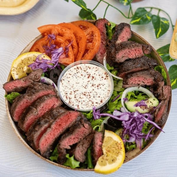 A steak salad with sliced tomatoes, red cabbage, lemon, and a side of white sauce.