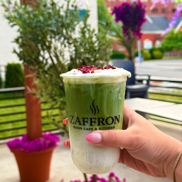 Hand holding a Zaffron Oasis Cafe & Kitchen matcha latte with rose petals on an outdoor patio.