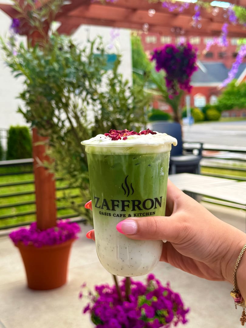 Hand holding a Zaffron Oasis Cafe & Kitchen matcha latte with rose petals on an outdoor patio.