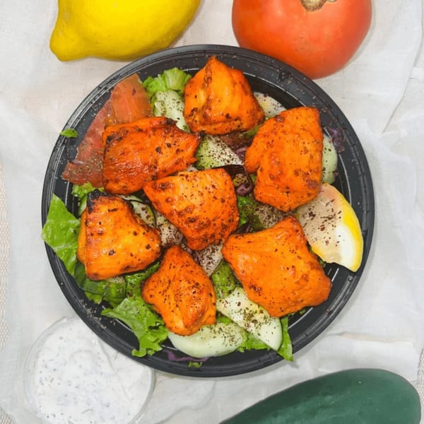 Chicken tikka salad with lettuce, cucumbers, tomatoes, and lemon wedges, served with a side of sauce.