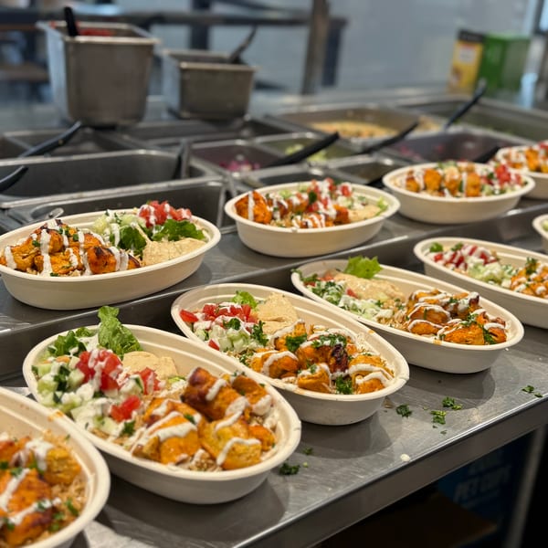 Multiple bowls of chicken and rice with salad and hummus, drizzled with white sauce on a counter.