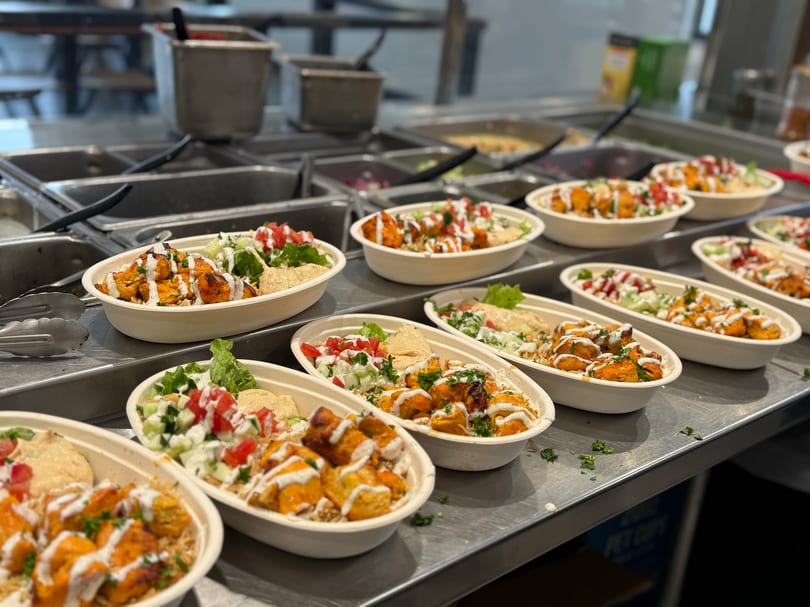 Multiple bowls of chicken and rice with salad and hummus, drizzled with white sauce on a counter.