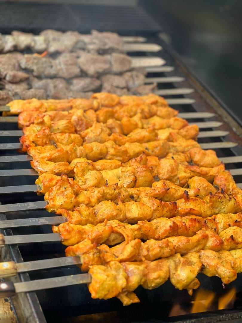 Chicken and beef kebabs cooking on a grill over flames, with smoke rising.