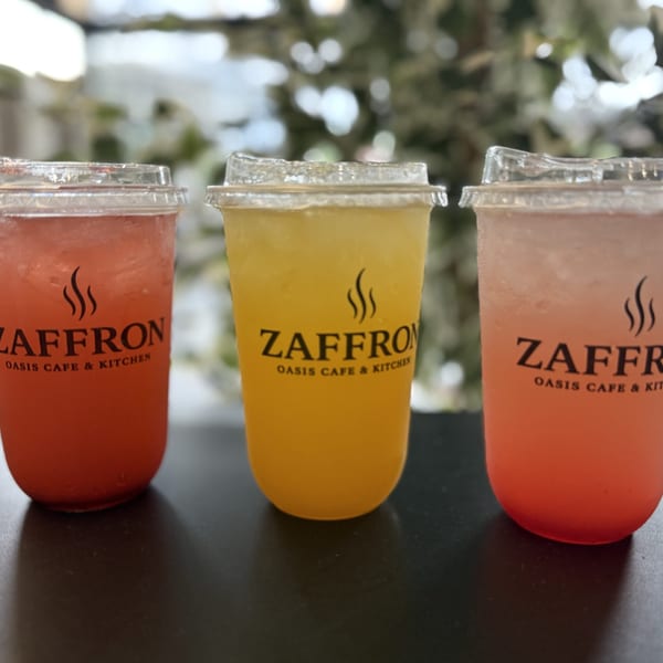 Three Zaffron Oasis Cafe & Kitchen drinks: red, yellow, and pink, on a dark table.
