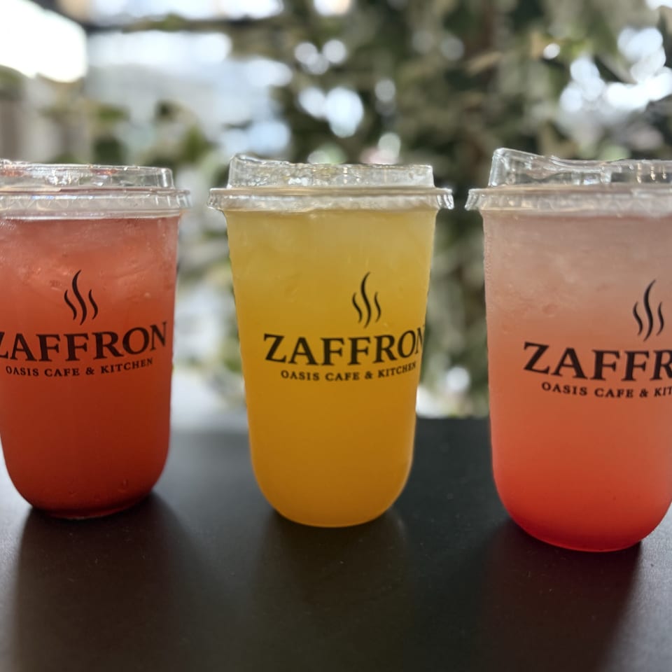 Three Zaffron Oasis Cafe & Kitchen drinks: red, yellow, and pink, on a dark table.