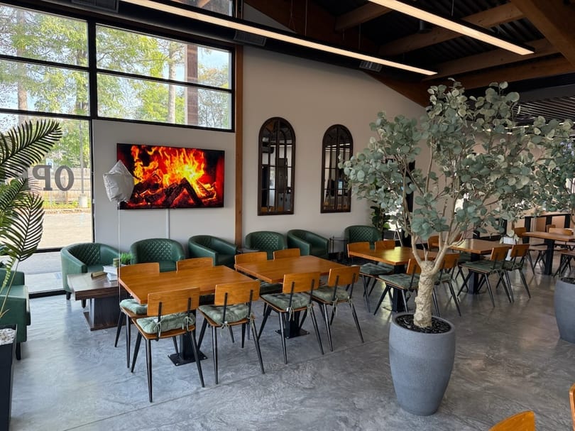 Modern restaurant interior with wooden tables, green chairs, large windows, and a digital fireplace.