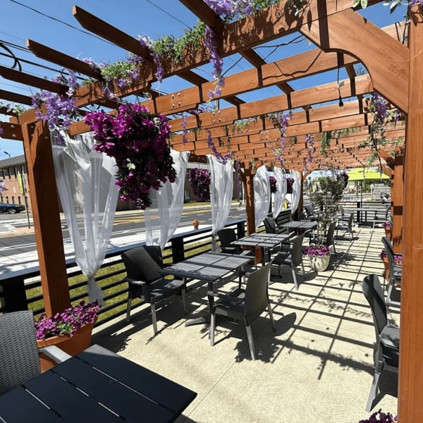 Outdoor patio with wooden pergola, purple flowers, white curtains, and dining tables with chairs.