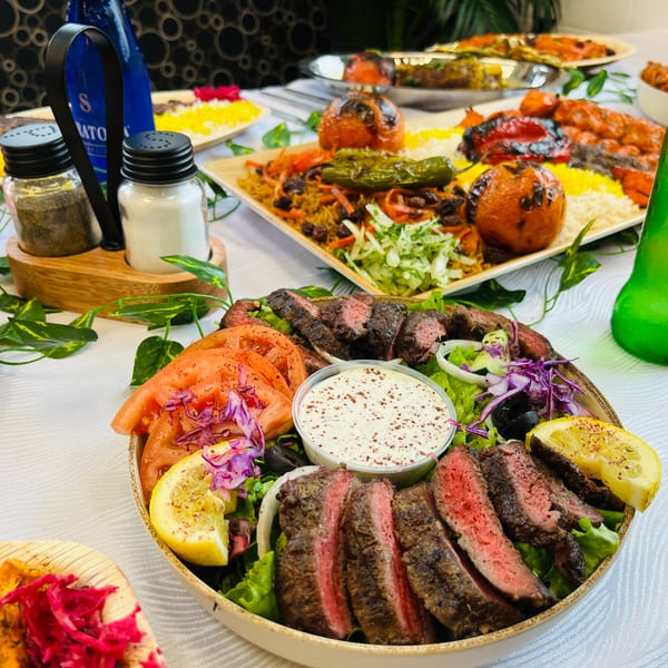 A table laden with various Middle Eastern dishes, including sliced steak, rice, grilled vegetables, and drinks.