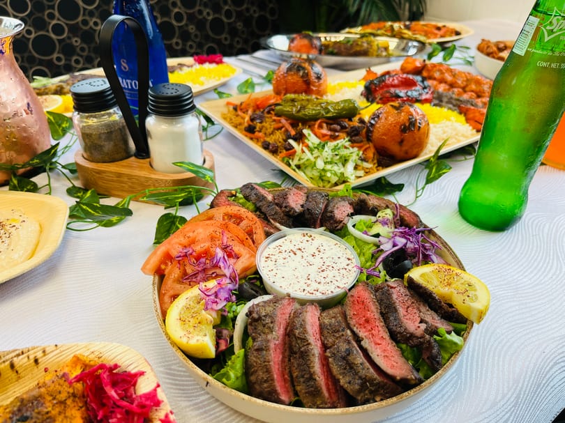 A table laden with various Middle Eastern dishes, including sliced steak, rice, grilled vegetables, and drinks.