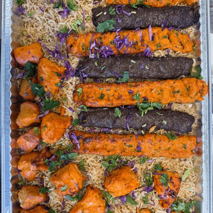 A catering tray filled with rice, orange chicken tikka, and dark seekh kebabs, garnished with herbs.