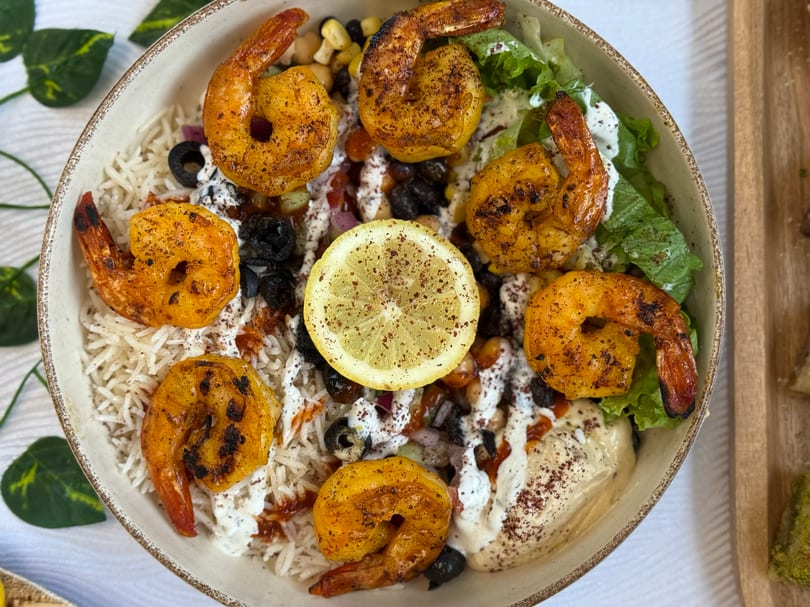 Overhead view of a shrimp bowl with rice, hummus, lettuce, olives, and a lemon slice.