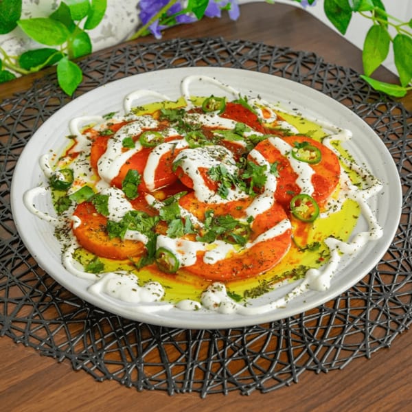 Sliced tomatoes drizzled with white sauce, olive oil, herbs, and green chili on a white plate.