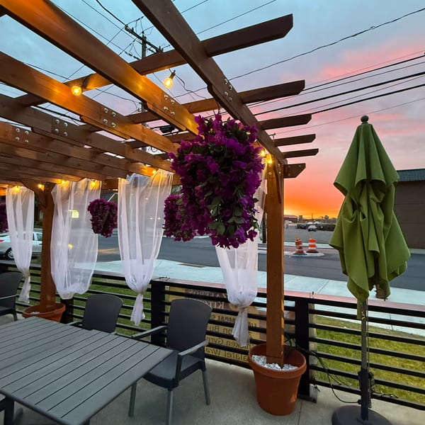 Outdoor patio with wooden pergola, string lights, purple flowers, and white curtains at sunset.