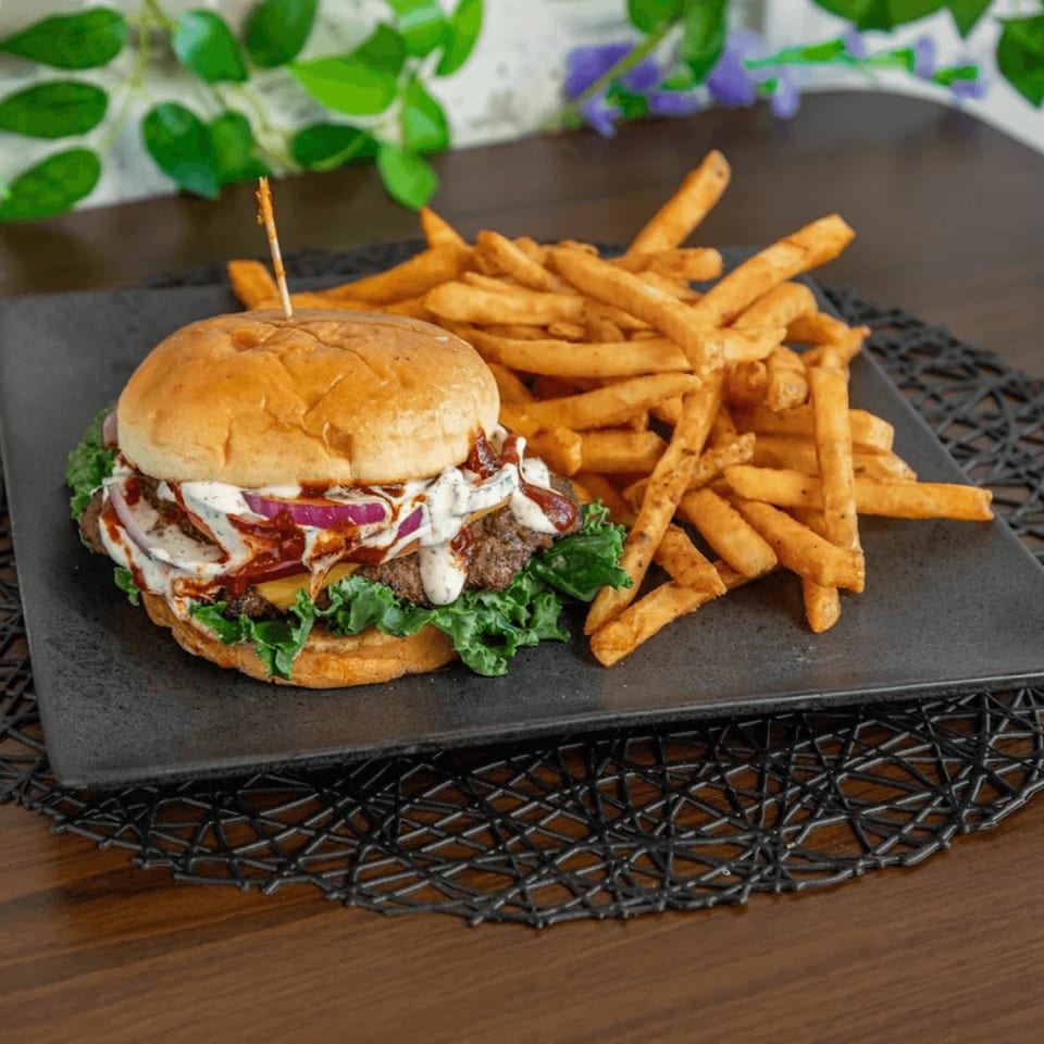A gourmet burger with red onions, sauce, and lettuce, served with golden french fries on a black plate.